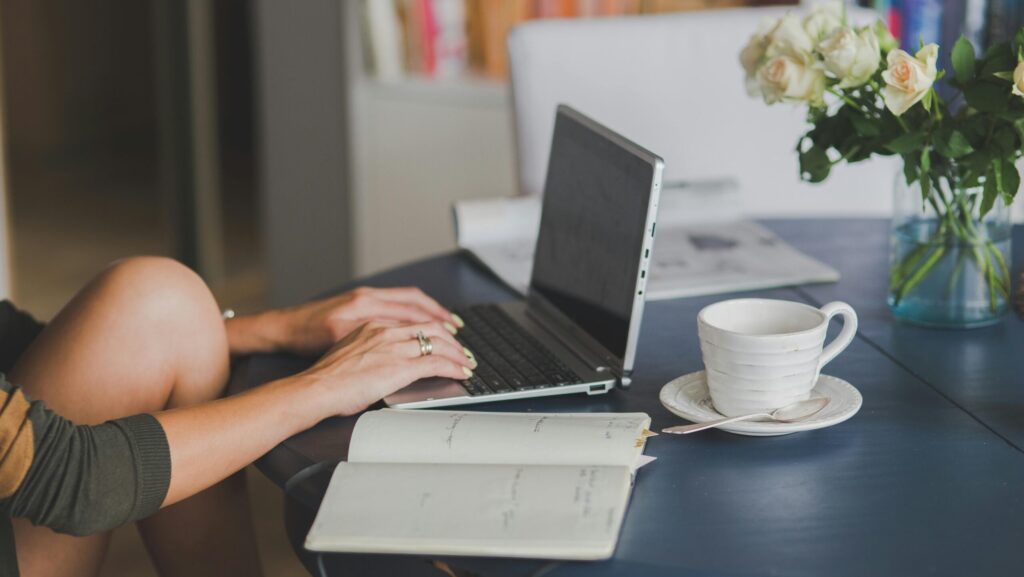 Female freelancer using laptop with coffee at home office desk, surrounded by roses and a planner.